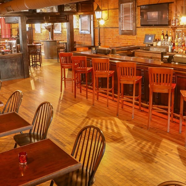 Warmly lit Firebird Tavern interior with a long wooden bar, stools, and dining tables on a hardwood floor.