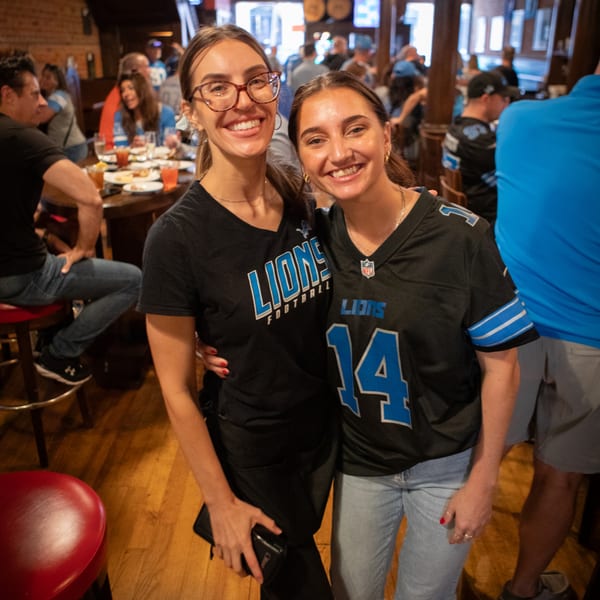 Two smiling women in Lions football jerseys pose in a lively Firebird Tavern bar.