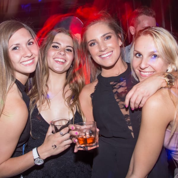 Four smiling women holding drinks and posing together in a dimly lit bar with red lighting.