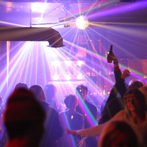 A disco ball casts colorful light beams over a lively crowd at Firebird Tavern, with one person holding a bottle up.