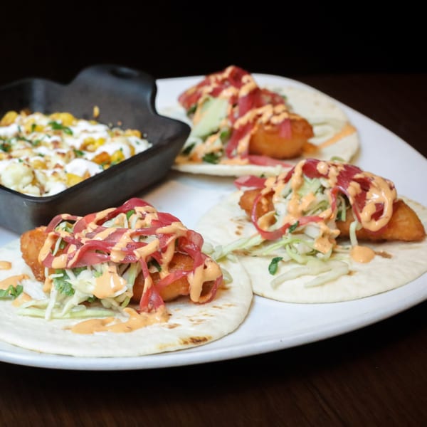 Three Firebird Tavern fish tacos with slaw, pickled onions, and sauce, alongside a side dish.