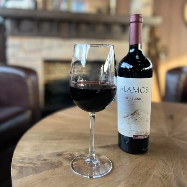 A glass of red wine and a bottle of Alamos Red Blend wine on a wooden table.