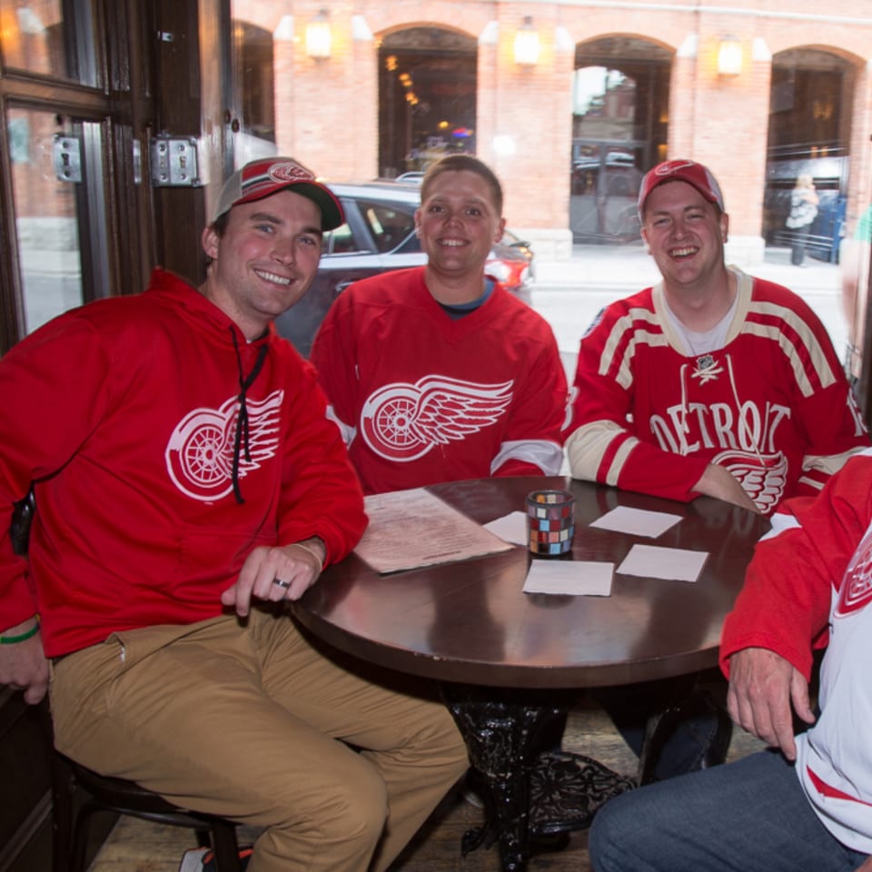 Four smiling Detroit Red Wings fans in red apparel gather around a restaurant table beside a window.