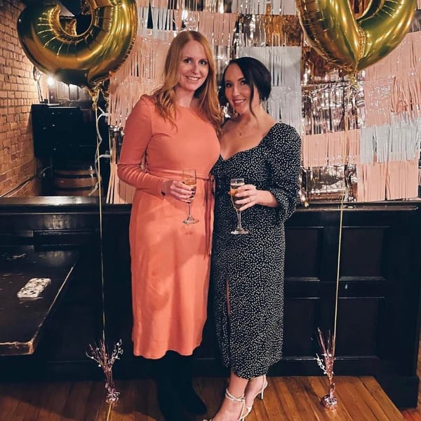 Two women holding champagne flutes, smiling in front of a festive backdrop with balloons at Firebird Tavern.