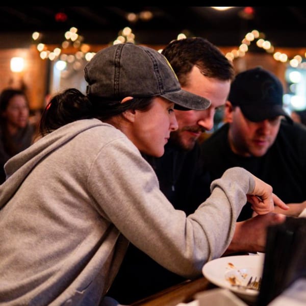 Three patrons at Firebird Tavern, one woman pointing at something on the table, with string lights in background.