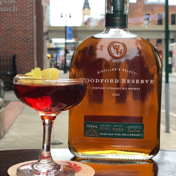 A Woodford Reserve whiskey bottle and a cocktail with lemon peel sit on a table by a window.