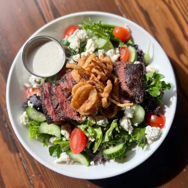 Steak salad with mixed greens, tomatoes, cucumbers, blue cheese, crispy onions, and a side of dressing.
