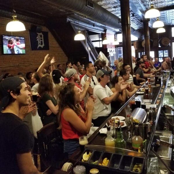 Patrons at Firebird Tavern bar watch a sports game on TV, clapping and cheering.