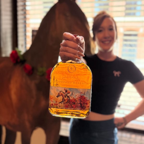 Smiling woman holding up a bottle of Woodford Reserve bourbon with a horse statue behind her.