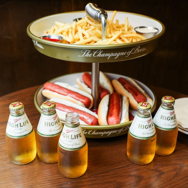 A two-tiered serving tray with fries and hot dogs, surrounded by six Miller High Life bottles.