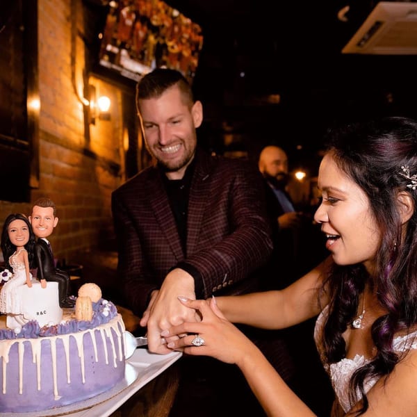 A smiling couple cuts a purple wedding cake with custom figurines at Firebird Tavern.