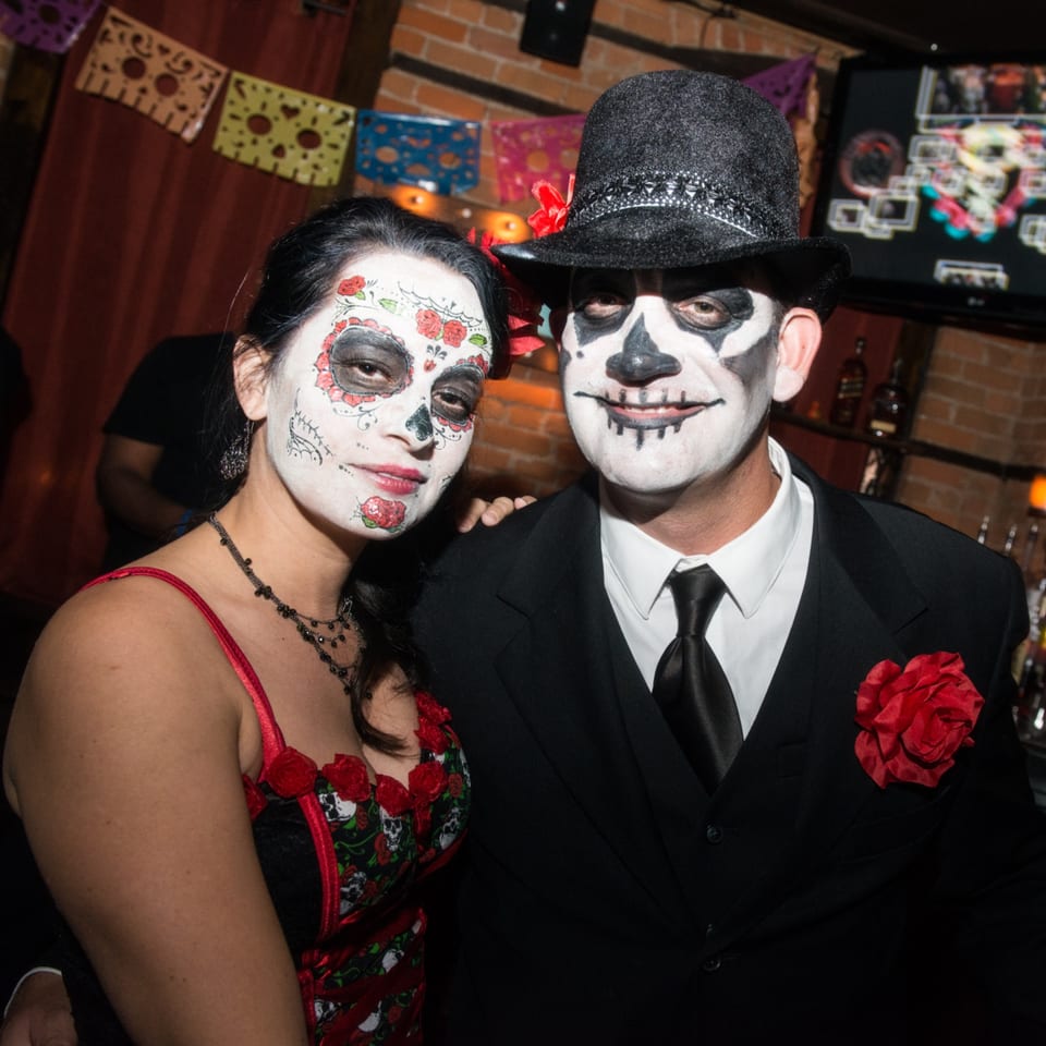 Two people in elaborate Día de los Muertos makeup pose in festive restaurant attire before colorful decorations and a brick wall.