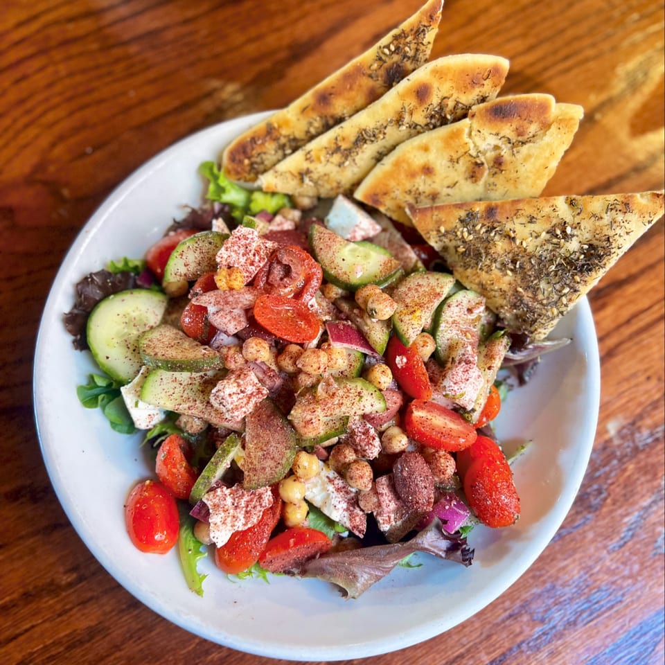 Greek-style salad with tomatoes, cucumbers, chickpeas, feta, olives, and herb-topped pita wedges on a white plate.
