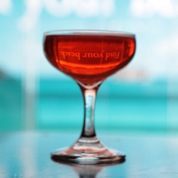 A red cocktail in a coupe glass with "Find your beach" reflected, against a blue background.