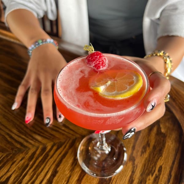 A person holds a pink cocktail garnished with a raspberry and lemon slice at Firebird Tavern.