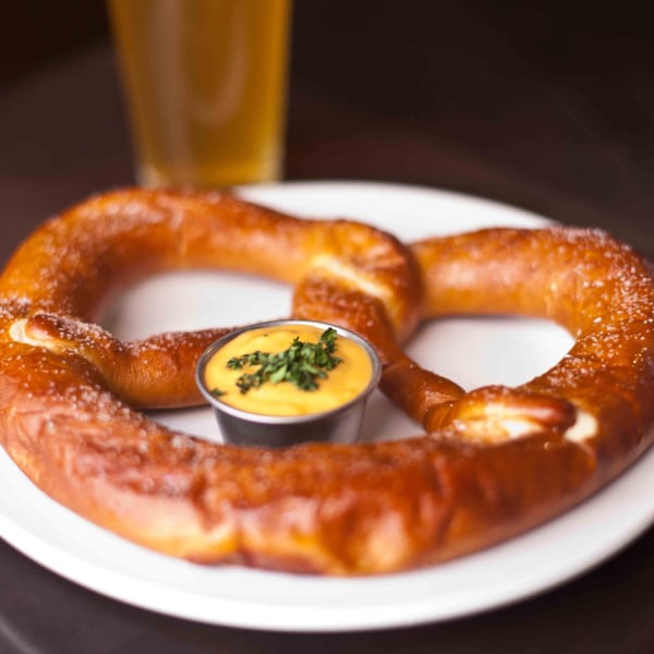 A large, salted pretzel with a side of cheese dip, garnished with parsley, and a beer.