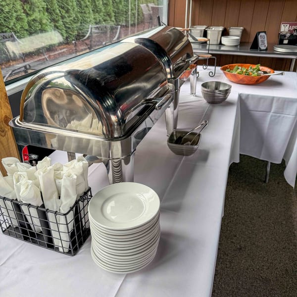A buffet setup with a chafing dish, stacked plates, rolled napkins, and a salad bowl.