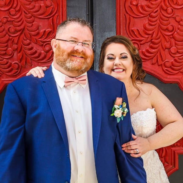 Smiling couple, a man in a blue suit and a woman in a white dress, against a red door.