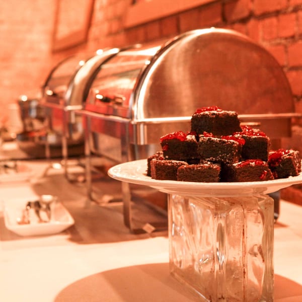 A plate of brownies with red jam and powdered sugar sits on a buffet table with chafing dishes.