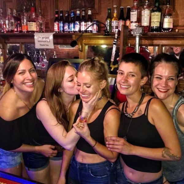 Five smiling women posing together in front of a bar with liquor bottles and beer.