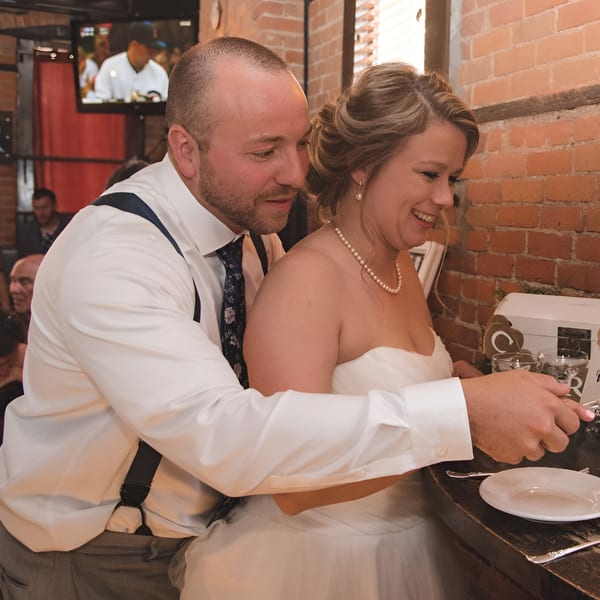 A smiling bride and groom cut a cake at Firebird Tavern, with brick walls and a TV in the background.