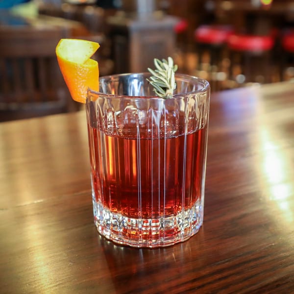 A Firebird Tavern cocktail, garnished with an orange peel and rosemary, sits on a wooden bar.