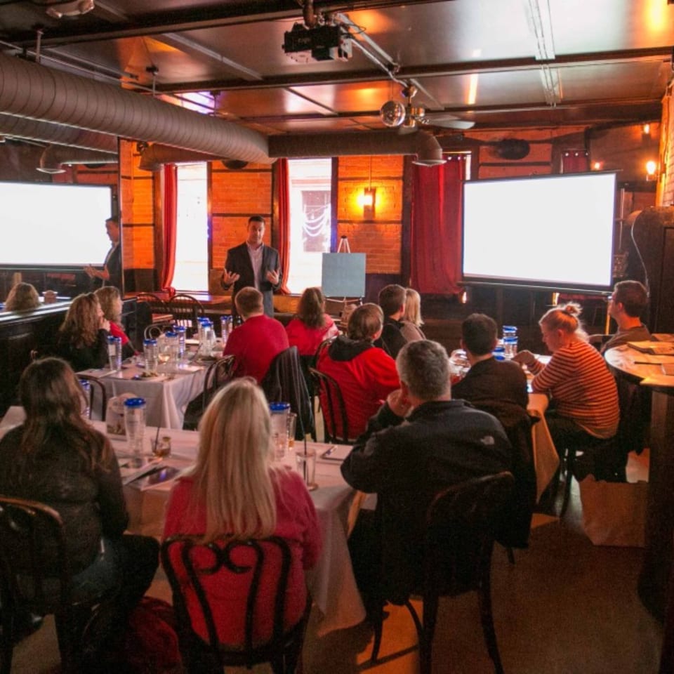 A speaker presents to an audience seated at tables in the Firebird Tavern dining room.
