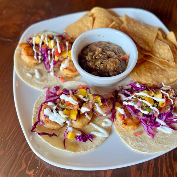 Three shrimp tacos with red cabbage, mango salsa, and crema, served with chips and salsa.