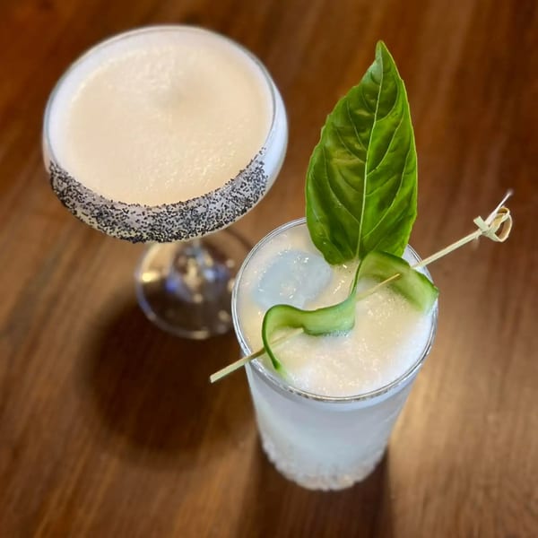 Two cocktails on a wooden table, one with a poppy seed rim, the other garnished with basil and cucumber.