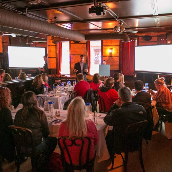 A man presenting to a group of people seated at tables in a dimly lit Firebird Tavern.